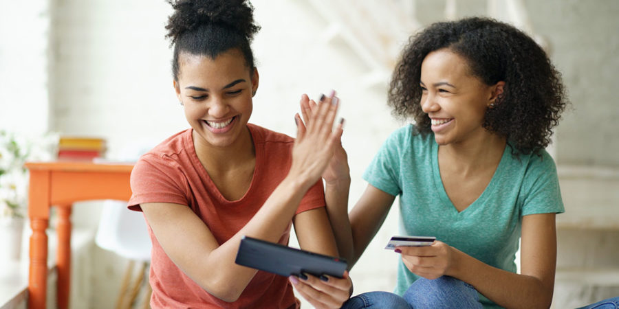Two cheerful mixed race curly girlfriends shopping online with tablet computer and credit card at home